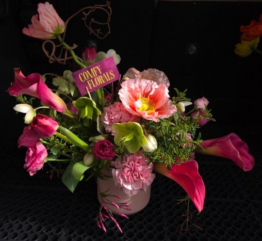 A bouquet of flowers feature pinks and reds and thin vines. A card for Comfy Florals is included in the arrangement.